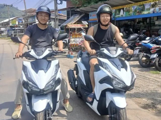นักท่องเที่ยวสองคนสวมหมวกกันน็อก นั่งบนมอเตอร์ไซค์เช่าสีขาวริมถนนข้างร้าน
